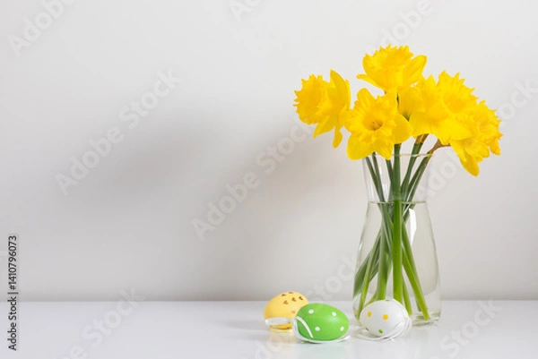 Fototapeta Easter background with yellow flowers in a glass vase and colored Easter eggs against a white wall, front view.