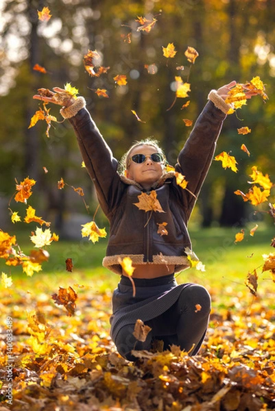 Fototapeta Beautiful young woman with autumn yellow leaves. Portrait of joyful woman playing with tree leaves in park during autumn. Happy girl wearing leather jacket and sunglasses and playing with leaves.