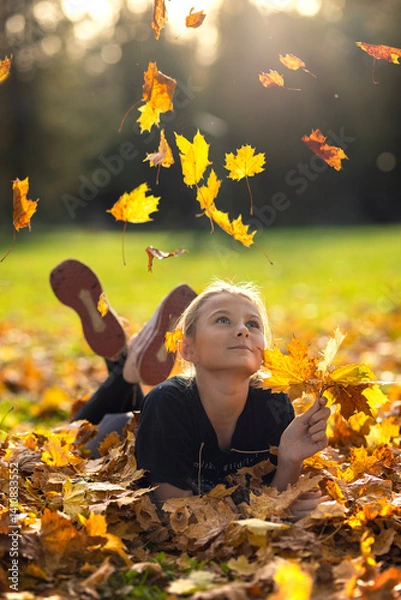 Fototapeta Beautiful young woman with autumn yellow leaves. Portrait of joyful woman playing with tree leaves in park during autumn. Happy girl wearing leather jacket and sunglasses and playing with leaves.