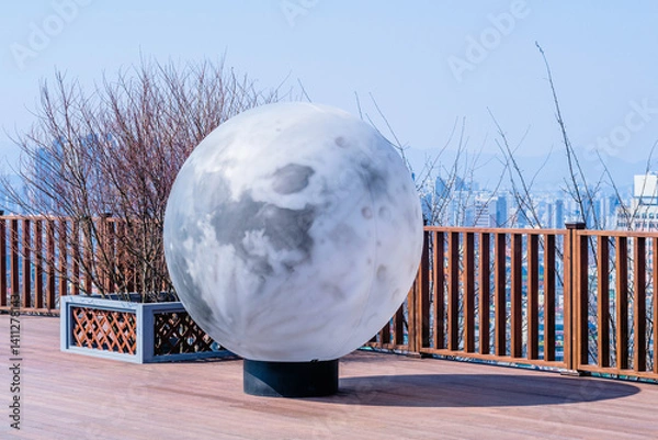 Fototapeta A large moon-like sculpture displayed on a wooden deck with city view.