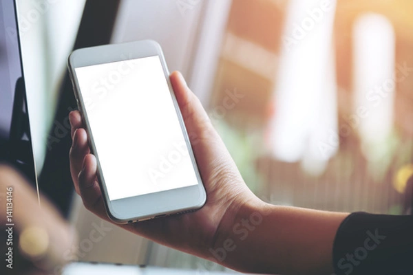 Fototapeta Mockup image of a hand holding white mobile phone and blank black screen with laptop in modern cafe
