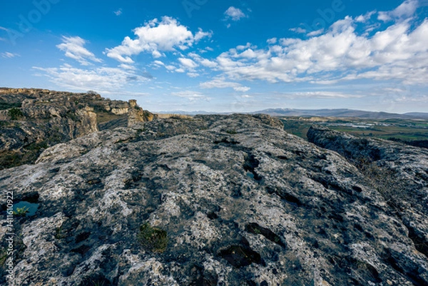 Fototapeta Wide view from a rocky plateau with dramatic textures, scattered clouds in a vivid blue sky, and distant green fields under mountain foothills.