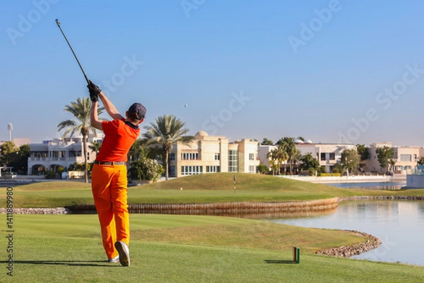 Obraz Golfer taking swing on luxury waterfront course, Male golfer in bright outfit swinging club on a high-end golf course near water.