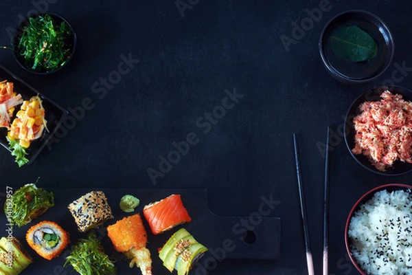 Fototapeta Top view of  assorted sushi rolls with salmon, avocado and wakame, crab meat, wakame salad, bowl of rice, green tea and chopsticks over black background. Copy space.