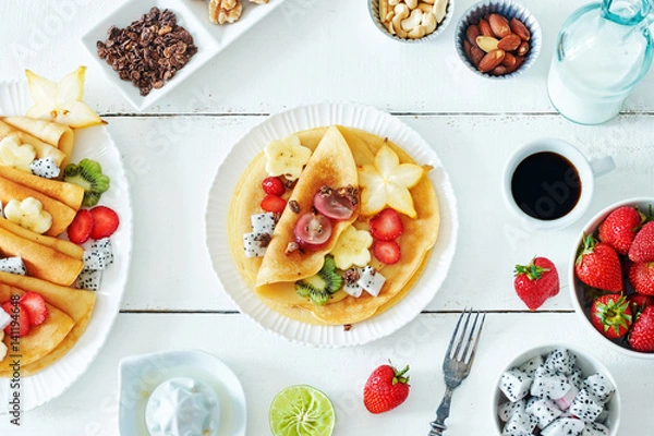 Fototapeta Top view of thin pancakes with starfruit, banana, kiwi, grape, strawberry and honey. Milk and coffee on a white table. Healthy breakfast with crepes. 