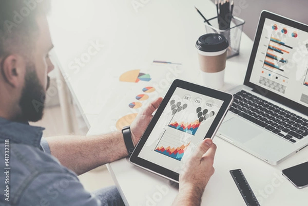 Fototapeta Tablet computer with graphs, diagrams and charts on screen in hands of young bearded businessman sitting at table
