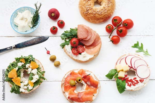 Fototapeta Assorted bagels with cream cheese, italian ham, cherry tomatoes, rocket salad, radish, cheddar, salmon and olives over white background. Copy space.