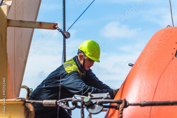 Obraz Portrait of the young Filipino officer, fully focused on his task during the inspection of the lifeboat and rescue boat. Dressed in a blue coverall and yellow helmet, he works with precision and care.