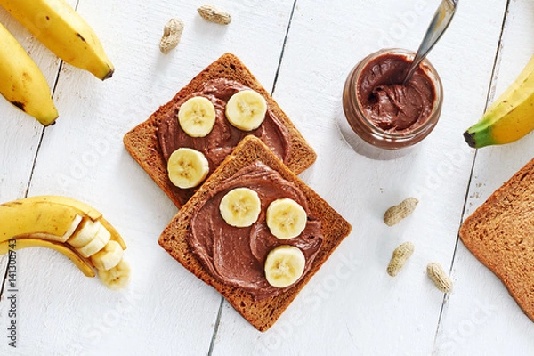 Fototapeta Portion of chocolate peanut butter sandwiches with rye bread and bananas over white background. Healthy snack with saturated fats. Top view.