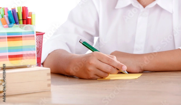 Fototapeta A boy student sitting at a desk and writing with a green pen on a yellow sheet of paper. 