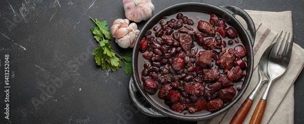 Obraz Feijoada, a traditional brazilian stew in a cast iron pot, accompanied by fresh garlic and parsley, ready to be served with a spoon and fork on a linen napkin, copy space