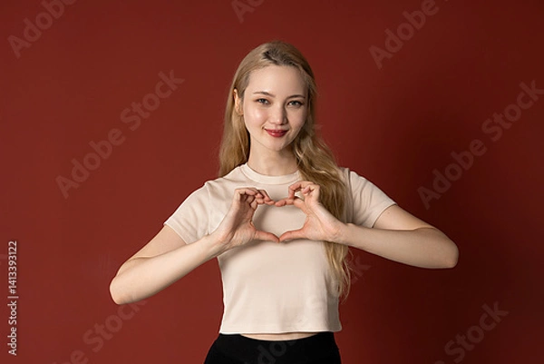 Obraz Portrait of smiling young asian girl with blond hair showing heart gesture with both hands and looking at camera, over red background. concept of romance and kindness. Copy space