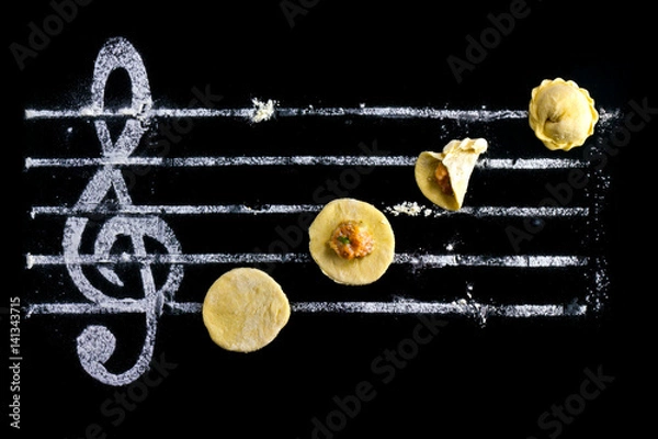 Fototapeta Ravioli - cooking music. Dough with minced meat on a black background and a treble clef from flour