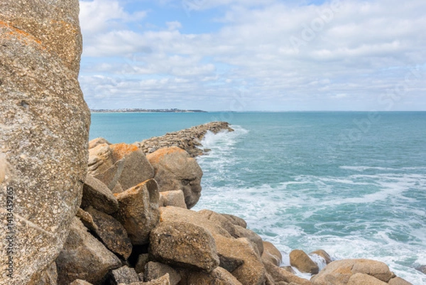 Obraz Waves hit a breakwater constructed of granite boulders with unbroken granite boulders in the foreground and the mainland in the distance under a cloudy sky at Granite Island in South Australia.