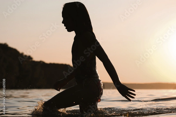 Fototapeta A silhouette of a person wading in water at sunset creates a peaceful, evocative scene. The water reflects the warm tones of the sky Tranquil ambiance