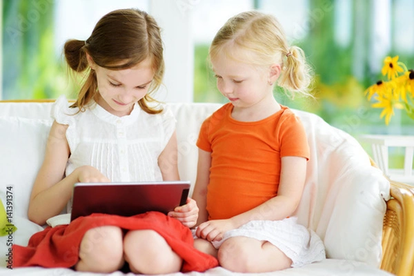 Fototapeta Two adorable little sisters playing with a digital tablet
