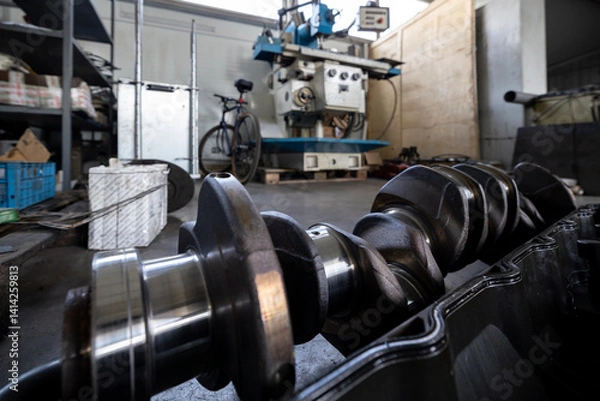Fototapeta A crankshaft and engine block lie on the floor of a repair shop, with a grinding machine standing ready in the background.