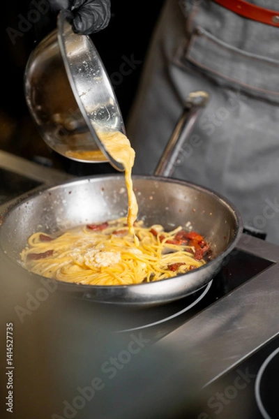 Fototapeta A chef in black gloves pours creamy sauce over cooked spaghetti and bacon in a stainless-steel skillet, demonstrating the art of culinary creation in a professional kitchen setting.