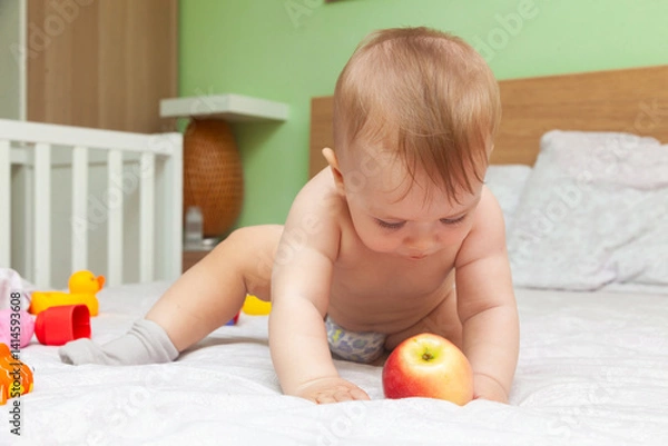 Obraz A baby on a bed tries to eat a red apple.