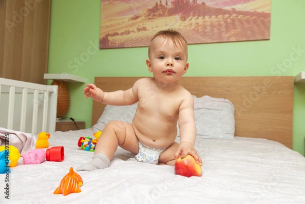 Obraz A baby on a bed tries to eat a red apple.
