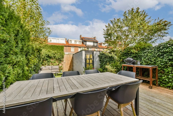 Obraz A stylish outdoor patio featuring a spacious wooden table surrounded by black chairs, set among lush greenery and blue skies. Ideal for gatherings and relaxation.