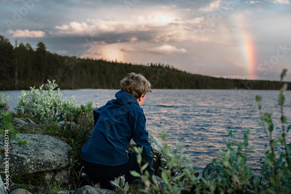 Fototapeta A boy sits crouched against rocks by the water, looks at the vibrant rainbow and sky as evening falls over the tranquil landscape of Dalarna, Sweden