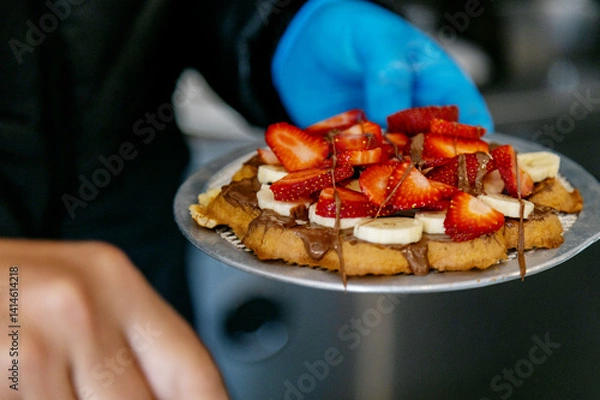 Obraz Freshly Made Waffle with Bananas, Strawberries and Chocolate, served on a metal plate. One person is holding the plate, wearing a blue glove