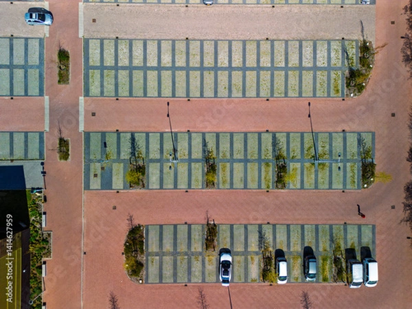 Fototapeta Aerial view of a nearly empty parking lot with a few parked cars, neat rows of green-grid spaces, brick walkways, and young trees casting long shadows in the sunlight.