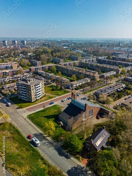 Fototapeta Aerial view of a residential neighborhood with a church at the center, surrounded by apartment buildings, green spaces, roads, and trees under a clear sky.