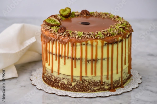 Fototapeta Pistachio Caramel Drip Cake featuring layers of moist cake drizzled with rich caramel, topped with chopped pistachios, and pecan halves with napkin on grey background side view of baked dessert