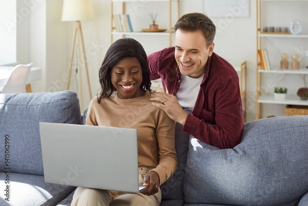 Fototapeta Young happy couple sitting on sofa in living room at home together watching funny video or using laptop for internet and social media. Happy man and African American woman resting on couch.