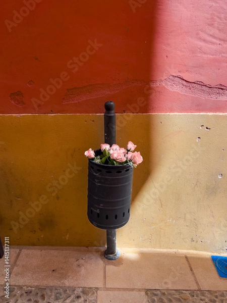 Fototapeta A black trash can filled with  thrown away Flower bouquet is affixed to a textured wall. The backdrop features warm hues of orange and yellow, creating a vibrant urban detail.
