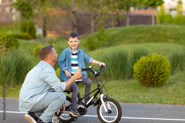 Fototapeta Father teaching son riding bike on the bike path. Father helping excited son to ride a bicycle in modern urban district. Happy child learning to ride cycle with his dad. Fathers day.