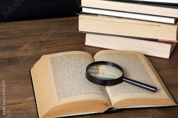 Fototapeta Close-up of an open book with a magnifying glass over it next to several stacked books.