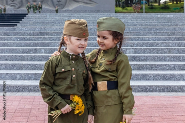 Fototapeta Patriotic education of children. Little girls with long braids in a military uniform, military cap, hold flowers in hands against the background of the memorial. Photo for the Victory Day on May 9
