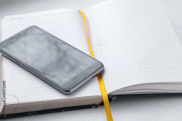 Fototapeta The cell phone is on open notebook with  yellow tab