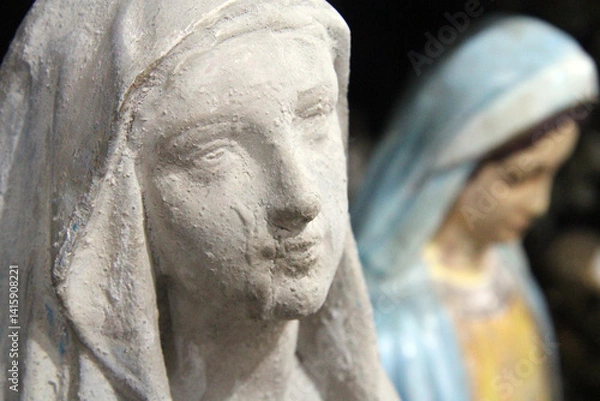 Obraz Close-up of a weathered statue of the Virgin Mary, with another colorful Madonna figure blurred in the background. A scene evoking devotion and sacred beauty.