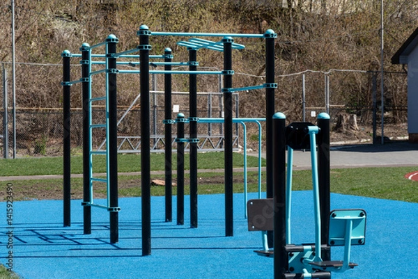 Fototapeta Various fitness stations are set up in a community park, featuring colorful equipment on a blue surface, inviting people to exercise outdoors in springtime