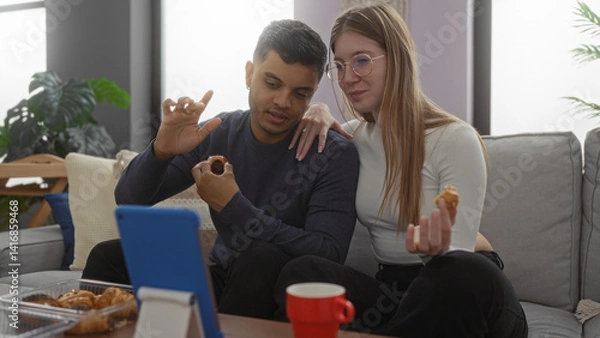 Obraz Couple enjoying a cozy video call together in their living room, sharing snacks and using a tablet while relaxing on the couch.