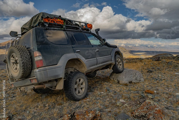 Fototapeta A family off-road touring car, on an off-road mud rubber, drove over a large stone, which is located on an elevated mountain. The mud rubber hit the object.