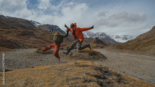 Fototapeta Two tourists in bright clothes make a jump in the mountains, and freeze in a funny pose. Behind them are the autumn mountains partially covered with snow.