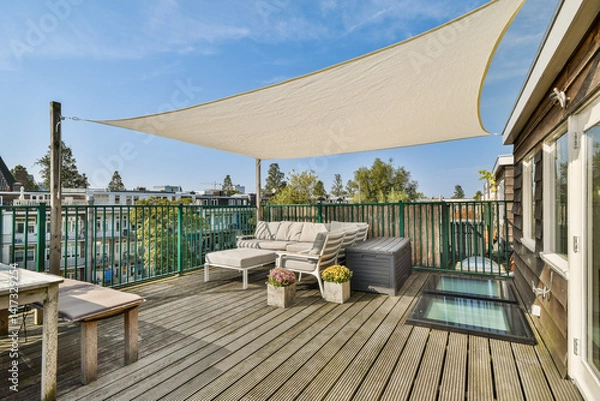 Fototapeta A stylish rooftop patio features comfortable seating, planters, and a protective shade sail, offering a relaxing outdoor space with a view.