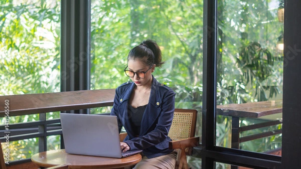 Fototapeta Businesswoman wearing glasses working on laptop in cafe, intently, working remote. A woman works on a computer in a coffee shop with a large mirror and plants.