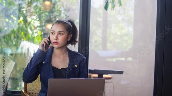 Obraz Asian businesswoman holding phone to ear and discussing business with partner and colleagues. Serious woman working remote in a cafe