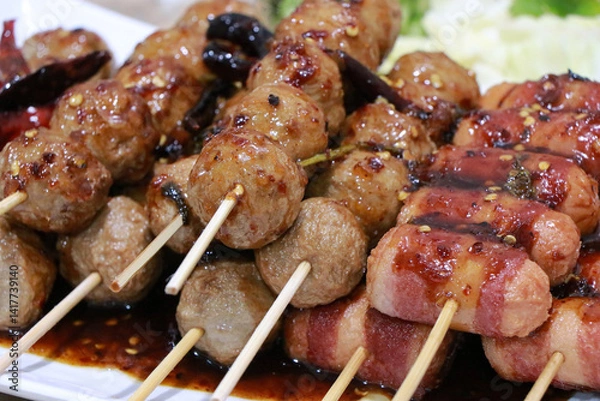 Fototapeta Grilled Meatballs. fish ball and sausage hotdog grilled in stick on tray, Fried pork meatballs skewers bamboo on a white plate with spicy sauce on a table, Thai street food.