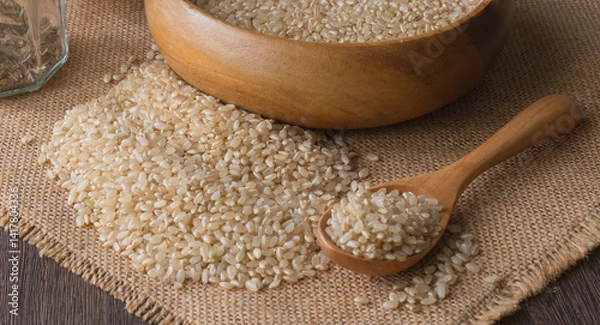 Fototapeta Background with brown rice grains in a wooden bowl and brown rice spread on a burlap cloth on a wooden table