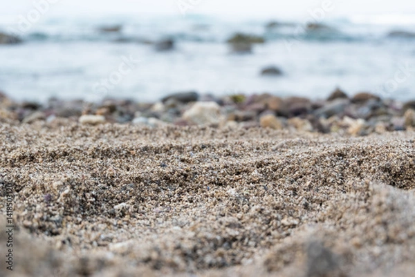 Obraz Macro shot of textured sand on a beach with ocean waves and blurred pebbles in the background. The detailed focus on the sandy surface highlights its natural granularity and coastal ambiance.