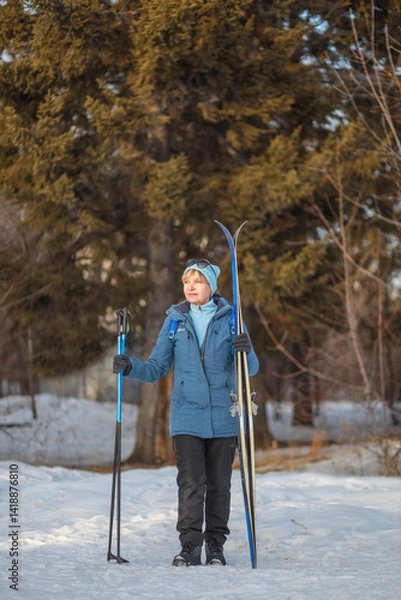 Fototapeta senior woman poses with her skis and poles in a tranquil winter park, embracing the joy of outdoor exercise. concept: active lifestyle, wellness, nature appreciation.