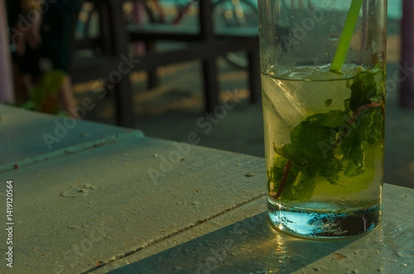 Fototapeta Ice cold mojito cocktail with mint leaves and a straw sitting on a table in an outdoor beach bar