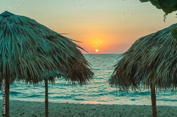 Fototapeta Beautiful sunset over the ocean with straw beach umbrellas and turquoise waters creating a relaxing vacation scene